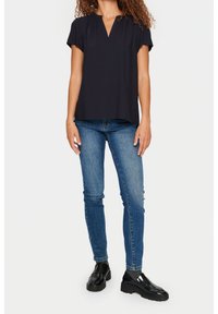 Donna che indossa una blusa nera a maniche corte, jeans skinny blu, calze grigie e mocassini neri spessi, in piedi contro uno sfondo bianco.