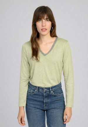 Jeune femme aux longs cheveux bruns portant un haut vert clair à manches longues avec un col en V et un jean bleu taille haute, sur un fond uni.