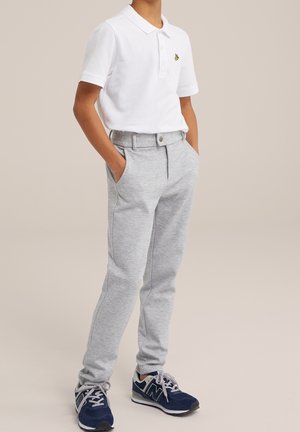 Niño viste una camiseta polo de manga corta blanca, pantalones ligeros grises con las manos en los bolsillos y zapatillas de deporte azul marino, de pie sobre un fondo liso.