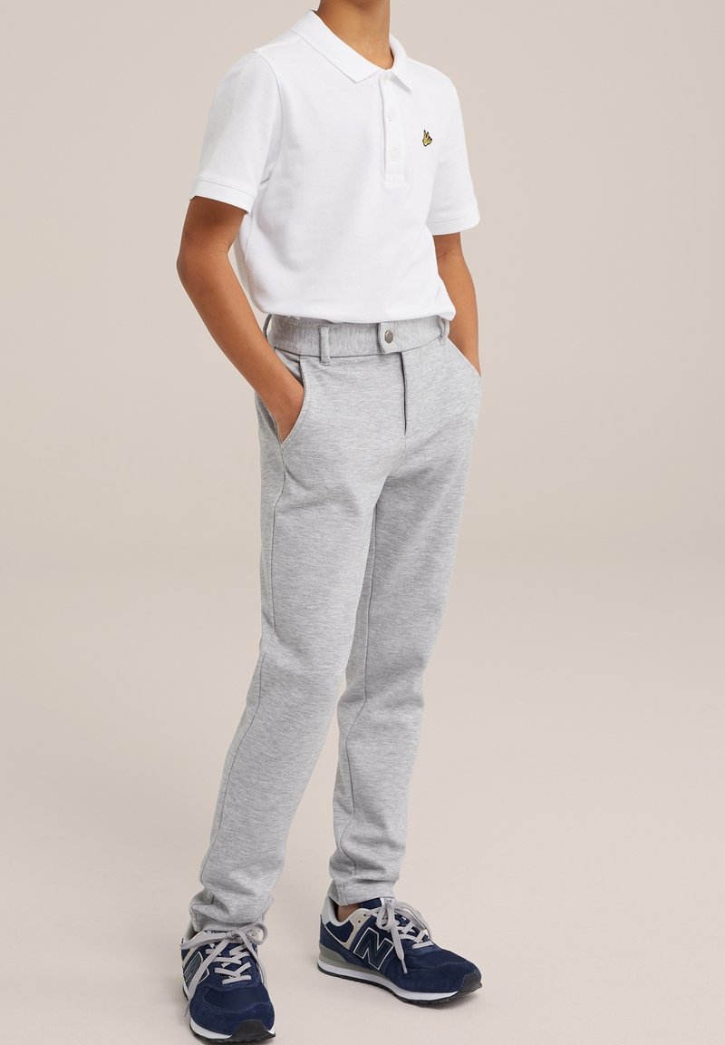 Dziecko ubrane w białą koszulkę polo z krótkim rękawem, jasnoszare spodnie z rękami w kieszeniach i granatowe sneakersy, stojące na jednolitym tle.