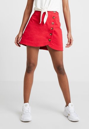 Vrouw draagt een rode, geknoopte wikkelrok, een wit geknoopt topje en witte sneakers, staand met gespreide benen tegen een effen achtergrond.