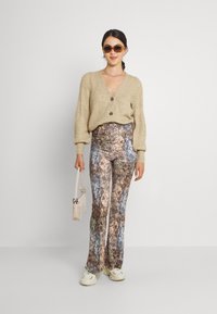 Beige gebreide cardigan met een V-hals, twee knopen. Geprinte flared broek in bruin- en blauwtinten, gecombineerd met witte schoenen en een zonnebril.