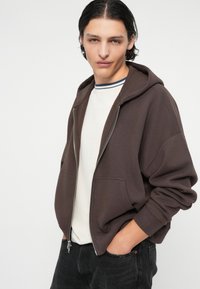 Veste à zip marron avec une grande capuche, poches avant, portée sur une chemise blanche avec des rayures bleues au col. Texture lisse, coupe décontractée.