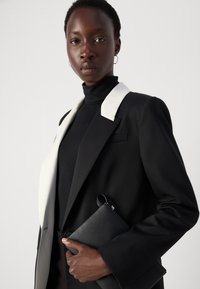 Black blazer with white lapels, layered over a black turtleneck. Holding a black clutch, featuring textured material and sleek design.