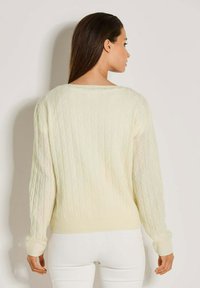 Frau mit langen dunklen Haaren in einem cremefarbenen Kabelstrickpullover und weißen Hosen, die mit dem Rücken zu einem schlichten hellen Hintergrund steht.