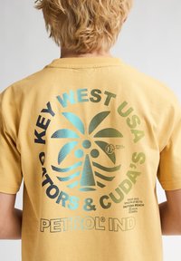 Gul bomulds t-shirt med et stort grafisk print i blå og sort, der viser palmer og tekst om Key West og Petrol Industries.