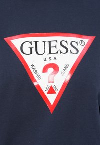 Rotes und weißes Dreieck-Logo auf dunkelblauem Stoff mit "GUESS U.S.A." in schwarzen Buchstaben und einem großen roten Fragezeichen in der Mitte.