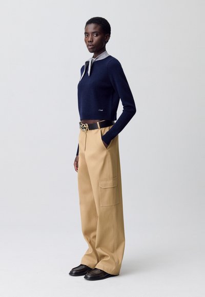 Pull léger bleu marine avec un col gris, associé à un pantalon large beige avec des poches latérales et une ceinture noire avec une boucle décorative.