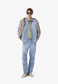 Un giovane posa con una giacca di denim chiara sopra una polo verde, indossando occhiali da sole e jeans azzurri chiari con una stampa paesaggistica.