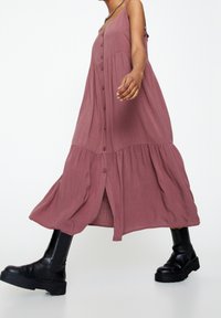 Vestido maxi en rosa empolvado, con botones frontales, diseño en capas y tirantes. Combinado con botines negros de suela gruesa.