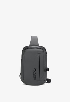 Zaino grigio inclinato realizzato in materiale resistente; presenta una tasca frontale con zip, una tracolla regolabile e un logo impresso sul lato.