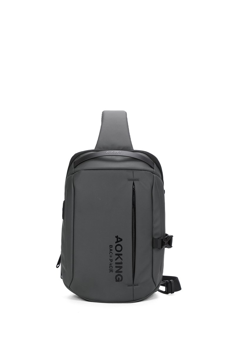 Zaino grigio inclinato realizzato in materiale resistente; presenta una tasca frontale con zip, una tracolla regolabile e un logo impresso sul lato.