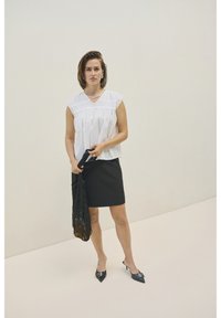Blouse blanche à manches courtes avec des détails froncés, assortie à une jupe crayon noire. Des chaussures à talons noirs et un châle en dentelle noire sont tenus.