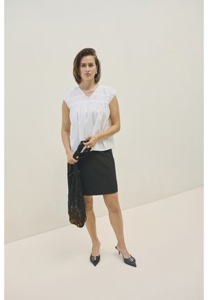 Blouse blanche à manches courtes avec des détails froncés, assortie à une jupe crayon noire. Des chaussures à talons noirs et un châle en dentelle noire sont tenus.