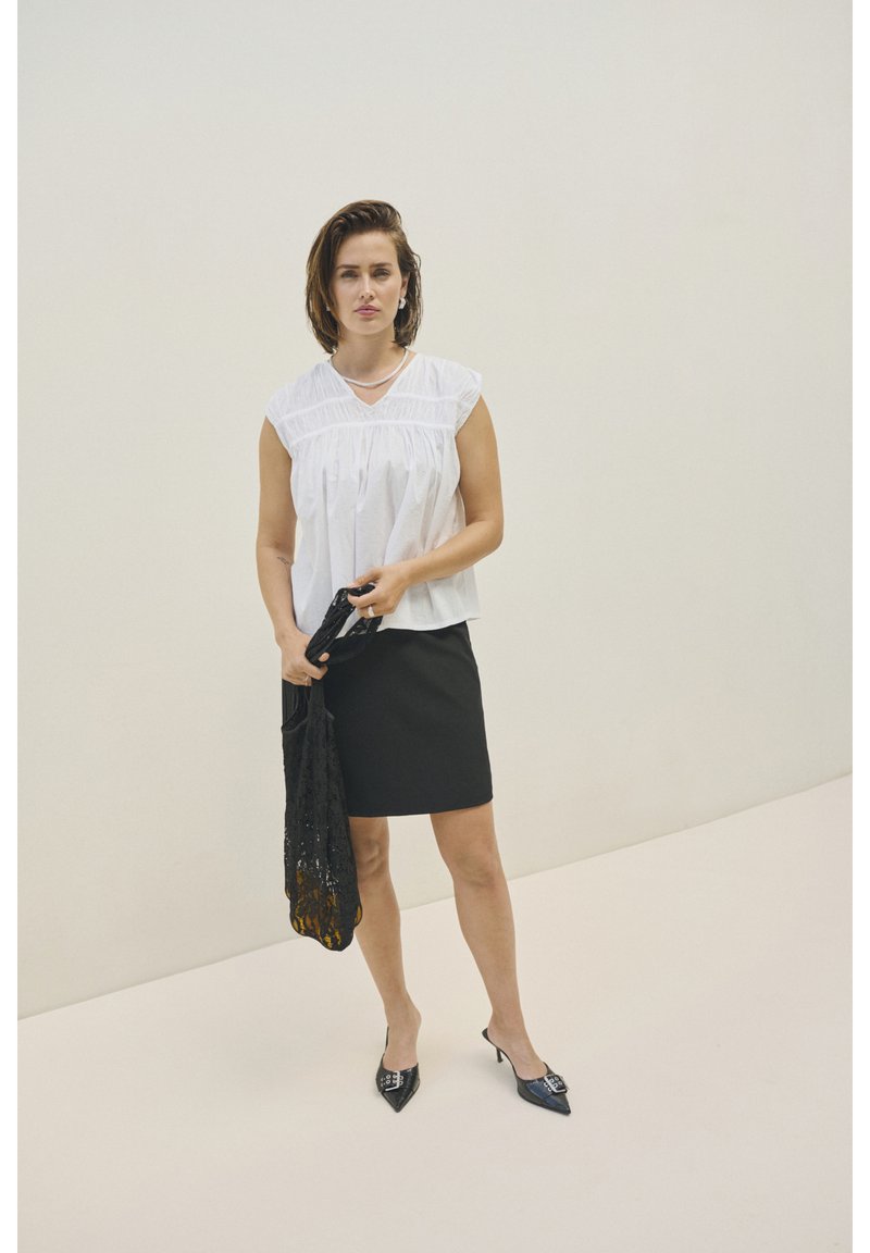 Blouse blanche à manches courtes avec des détails froncés, assortie à une jupe crayon noire. Des chaussures à talons noirs et un châle en dentelle noire sont tenus.