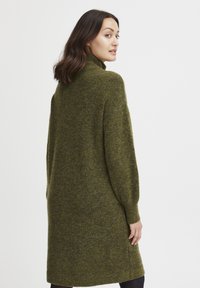 Vestito longo verde a maglia, morbido e ampio, con colletto alto, polsini a coste e un tessuto texture. Arriva a metà coscia.