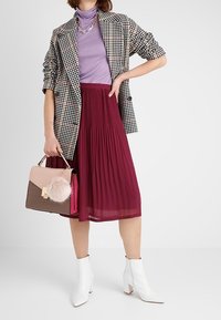 Kobieta w marynarce w kratę houndstooth, fioletowym golfie, bordowej spódnicy plisowanej, białych botkach za kostkę, trzymająca torebkę z puszystym różowym breloczkiem.