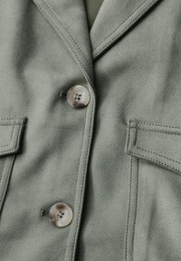 Green fabric blazer featuring a tailored design, two patch pockets, and a large, iridescent button at the front. Textured finish.