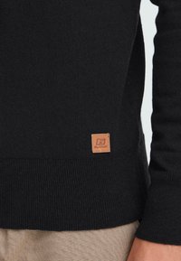 Suéter negro de punto con una pequeña etiqueta de cuero marrón etiquetada "Blend" cerca del dobladillo acanalado, llevado sobre pantalones beige texturizados.