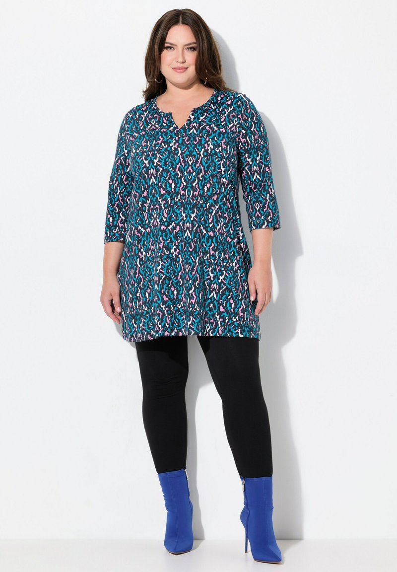 Multicolore tuniek met een teal, roze en lavendel patroon; voorzien van driekwart mouwen en een V-hals ontwerp, in combinatie met zwarte leggings en blauwe enkellaarsjes.