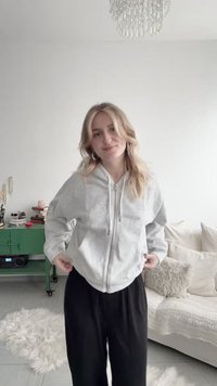 Νεαρή γυναίκα που προσαρμόζει το ανοιχτό γκρι φούτερ με φερμουάρ, στέκεται σε ένα ζεστό σαλόνι με λευκό καναπέ, αφράτα μαξιλάρια και πράσινο πλαϊνό τραπεζάκι.