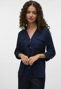 Blusa blu navy con scollatura a V, maniche lunghe arrotolate fino ai gomiti, due tasche sul petto e bottoni color oro. Tessuto dalla texture liscia.