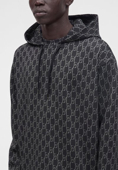 Sweat à capuche noir avec un motif géométrique en lignes blanches couvrant le tissu, porté par une personne à la peau foncée, visible des épaules jusqu'aux lèvres.