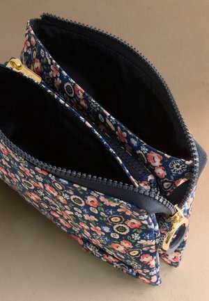 Pungă din material floral albastru marin cu fermoar auriu, compartimente duble și căptușeală interioară neagră, plasată în unghi pe o suprafață bej.