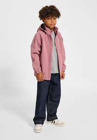 Rosa leichte Jacke mit Kapuze, elastischen Bündchen, getragen über einem grauen Shirt, kombiniert mit dunkelblauen Jeans und weißen Sneakers.
