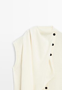Gilet en tricot sans manches de couleur crème avec une écharpe assortie portée sur l'épaule, comportant trois boutons noirs à l'avant.