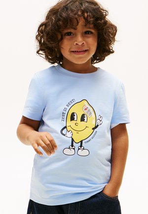 Barn med krøllet hår iført en lyseblå skjorte med en tegneseriecitron, der holder op med et fredstegn og teksten "Zest is Best!"