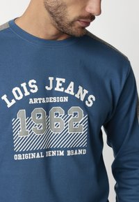 Sudadera azul con acentos en gris en los hombros, que presenta texto en blanco: "LOIS JEANS ART&DESIGN 1962 MARCA DE DENIM ORIGINAL" y un patrón a rayas.