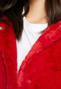 Rød faux fur jakke med en blød, tekstureret overflade og trykknapdetalje, båret over en hvid skjorte.