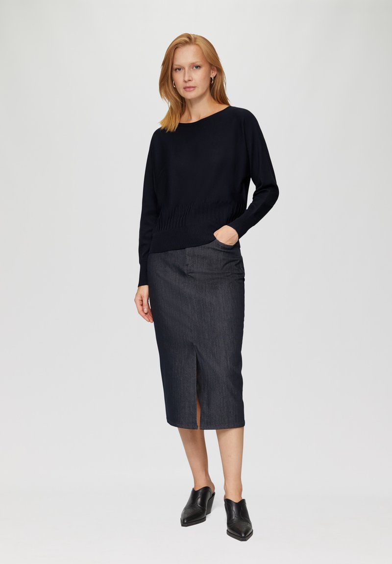 Black knitted sweater with a relaxed fit and ribbed details, paired with a dark denim pencil skirt featuring a front slit. Black heeled mules complete the look.