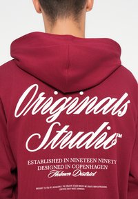Burgundi kapuutsärk, mille seljal on suur valge graafiline tekst, kus on silmapaistvalt kirjas "Originals Studio". Tekst sisaldab brändi üksikasju.