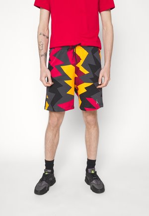 Person iført farverige zigzag-mønstrede shorts i rød, gul, sort og grå med sorte sneakers og en rød skjorte, stående mod en hvid baggrund.