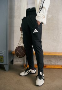 Schwarze Sporthosen mit einem Kordelzugbund, die weiße Adidas-Streifen und ein Sportlogo aufweisen, kombiniert mit weißen und schwarzen Schuhen und einem Basketball in der Hand.