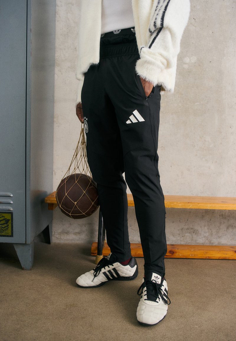 Schwarze Sporthosen mit einem Kordelzugbund, die weiße Adidas-Streifen und ein Sportlogo aufweisen, kombiniert mit weißen und schwarzen Schuhen und einem Basketball in der Hand.