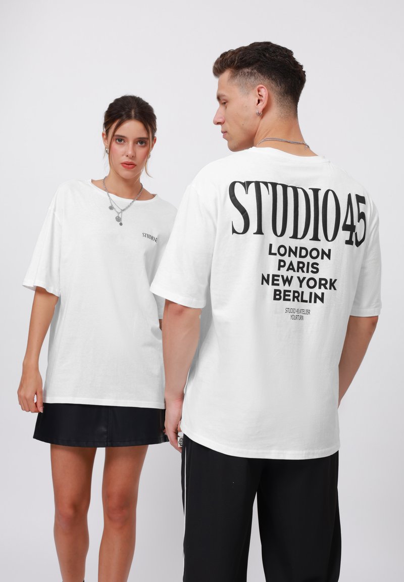 Camiseta oversized blanca con la leyenda "STUDIO 45" y nombres de ciudades en impresión negra en la espalda. Hecha de algodón suave, con un corte holgado y mangas cortas.