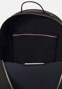 Interno di uno zaino nero con una consistenza liscia. Presenta un compartimento con cerniera e una tasca con una striscia rossa, bianca e blu.