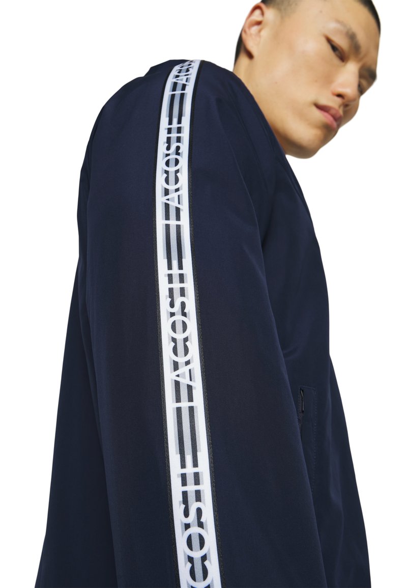 Marineblaues Jackett mit weißem Logostreifen entlang des Ärmels. Verfügt über eine glatte Oberfläche und ein modernes, lockeres Design mit einer Reißverschlusstasche.