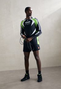 Schwarzes Sportshirt mit grünen und weißen Akzenten, Shorts und schwarzen Schuhen. Das Shirt hat ein Logo und Netzdetails. Hält ein Springseil.