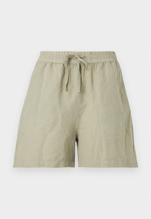 Sötét hajú fiatal nő, aki bézs rövid ujjú felsőt, világoszöld, húzózsinóros rövidnadrágot és krémszínű hegyes orrú lapos cipőt visel fehér háttér előtt.