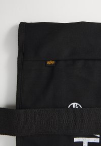 Bolsa mensajera negra de lienzo con solapa, que presenta una pequeña etiqueta de logo y una correa texturizada. El material es duradero y resistente.