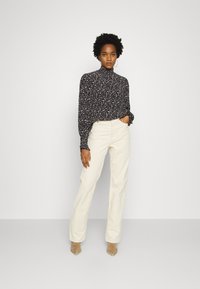 Blusa de flores de manga longa com gola alta, fundo escuro e flores multicoloridas, combinada com calças de cintura alta em bege claro e sapatos nude.