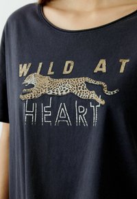 Black cotton t-shirt featuring a leopard graphic, the text "WILD AT HEART" in metallic gold and silver accents, with a relaxed neckline.