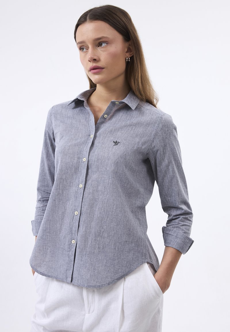 Camicia button-up in blu chiaro/grigio, maniche lunghe, colletto, texture morbida e un sottile logo di un uccello sul lato sinistro del petto. Abbinata a pantaloni bianchi.
