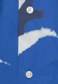 Camicia blu a fantasia con una texture di tessuto liscia, caratterizzata da un design bianco, un bottone metallico circolare e un asola cucita in blu.