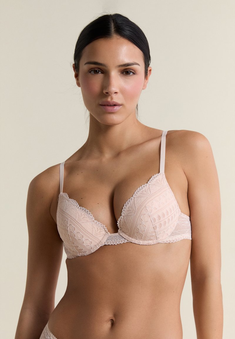 Donna con capelli scuri in un reggiseno di pizzo rosa chiaro, rivolta verso l'obiettivo su uno sfondo beige semplice.