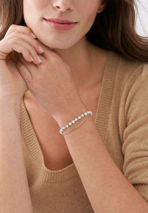 Perlenarmband mit blauen und weißen Steinen, goldene Kettenakzent; getragen an einem Handgelenk mit heller Haut, vor einem Hintergrund aus weichem Stoff.